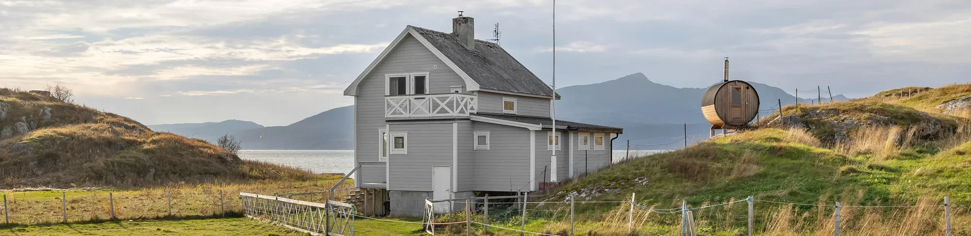 Weisses Holzhaus mit Fass-Sauna am Hang, dahinter Fjord und Berge